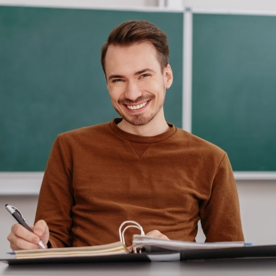 Happy male student with a confident beaming smile sitting working on paperwork in the classroom in front of a chalkboard